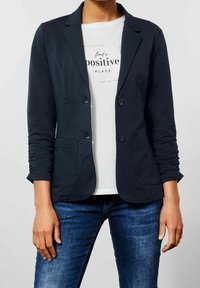 Námořnický blazer s přizpůsobeným střihem, dvěma předními kapsami a zapínáním na knoflíky, přes bílou košili se textem, v kombinaci s tmavými džíny.