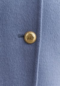 Tessuto di feltro blu con una texture morbida, caratterizzato da un bottone dorato con un design intricato, posizionato lungo una cucitura per la chiusura.