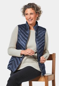 Marineblauer gesteppter Weste mit hohem Kragen, aus glänzendem Synthetikmaterial, über einem hellgrauen Strickpullover, kombiniert mit schwarzen Hosen.