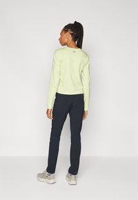 Lichtgroen langemouwen top, aangesloten ontwerp, gecombineerd met donkere broek. Model draagt grijze schoenen met paarse accenten. Eenvoudige achtergrond.