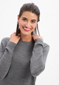 Pull en tricot gris avec un col rond et des manches longues, présentant une texture douce. Le modèle a les mains près des épaules, affichant un sourire subtil.