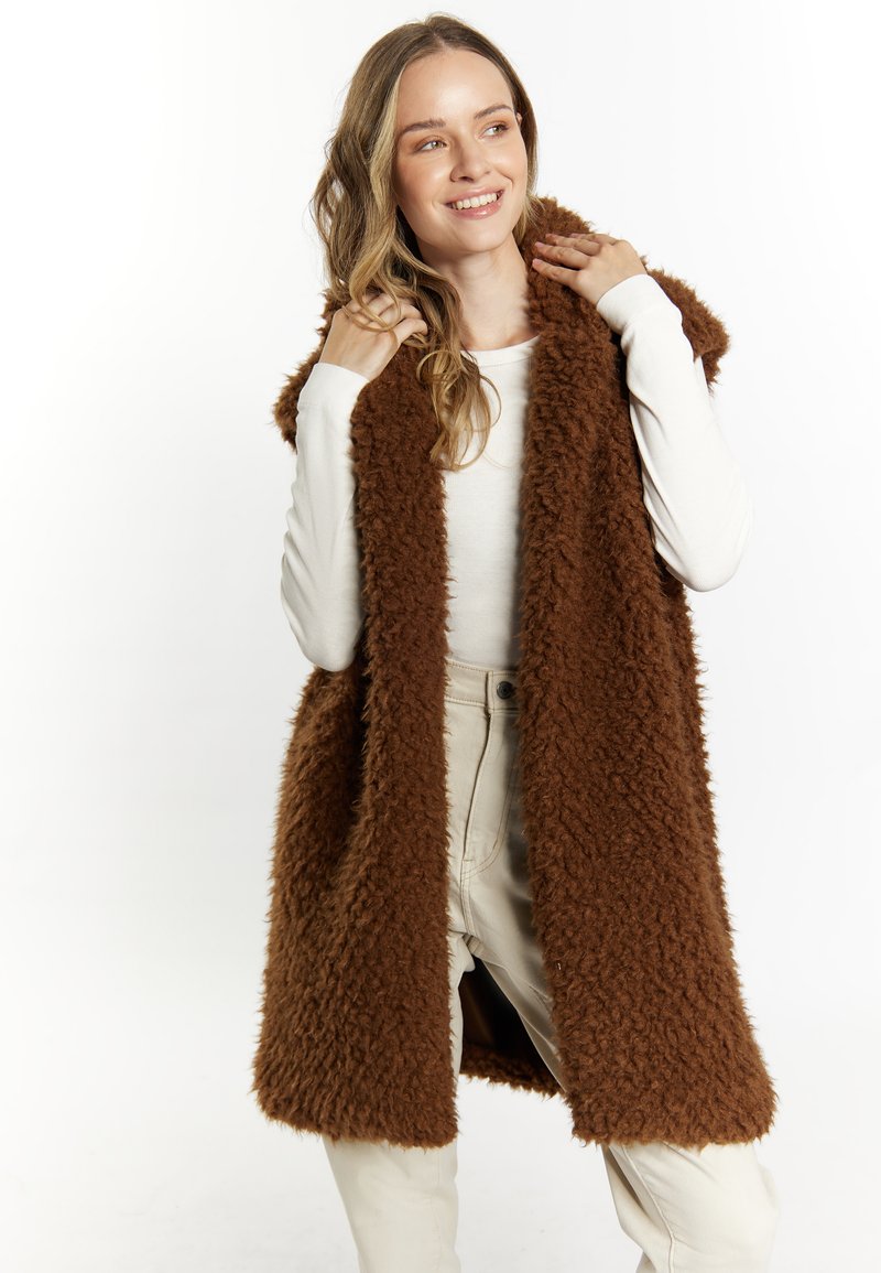 Bruine nepbont vest met een zachte, harige textuur. Open voorkant zonder sluitingen, gedragen over een aansluitend wit langarmtop en lichtbeige broek.