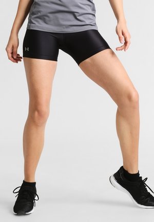 Athlete wearing black Under Armour compression shorts, gray top, and black running shoes standing with legs apart on a gray background.