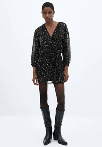 Abito nero a forma di sarong con maniche lunghe e trasparenti, caratterizzato da un motivo a macchie bianche e una vita aderente. Indossato con stivali neri fino al ginocchio.