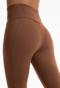 Leggings marron taille haute en tissu extensible, avec une texture lisse et une silhouette ajustée, mettant en avant des détails de contour.
