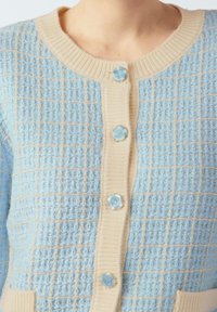Jasnoniebieski dzianinowy kardigan z fakturowanym wzorem w kratę, kremowym prążkowanym kołnierzem i listwą na guziki, z okrągłymi niebieskimi guzikami w kwiaty.