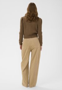 Ženska z rjavimi valovitimi lasmi, oblečena v rebrast rjav pulover in široke bež kord flanelne hlače, stoji pred enostavno belo ozadjem.