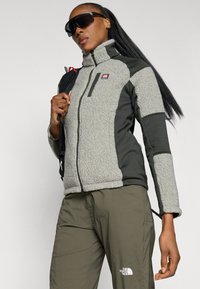 Chaqueta de forro polar gris con detalles en verde oscuro, cuello alto, bolsillo en el pecho con cremallera, combinada con pantalones caqui que presentan un logo. Gafas de sol negras.