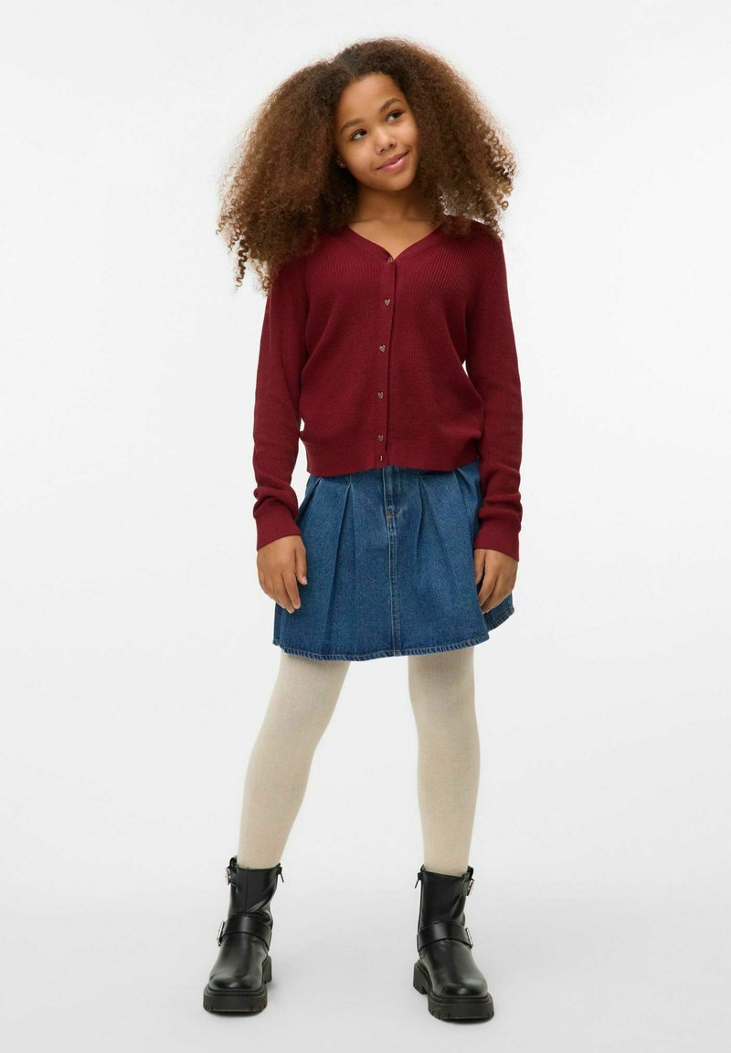 Tamsiai raudonas banguotas kardiganas su auksiniais širdelės mygtukais, derinamas su klostuota džinsine sijonu, smėlio spalvos pėdkelnėmis ir juodais kulkšninių batais.