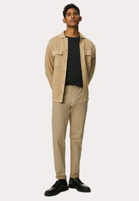 Beige skjortejakke over en sort skjorte, parret med lysebeige bukser og sorte sko. Glatte teksturer og standard pasform hele vejen igennem.