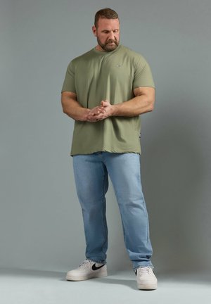Mężczyzna z krótkimi włosami i brodą stoi, patrząc w dół, ubrany w oliwkowy t-shirt, jasnoniebieskie dżinsy i białe sneakersy na tle jednolitego tła.