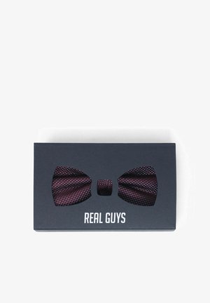 Dunkellila Fliege mit weißen Punkten, präsentiert in einer schwarzen Schachtel mit der Aufschrift "REAL GUYS" auf weißem Hintergrund.