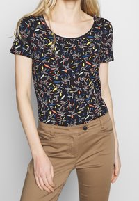 Black short-sleeve top with a colorful leaf pattern, featuring a scoop neckline. Paired with light brown fitted pants.