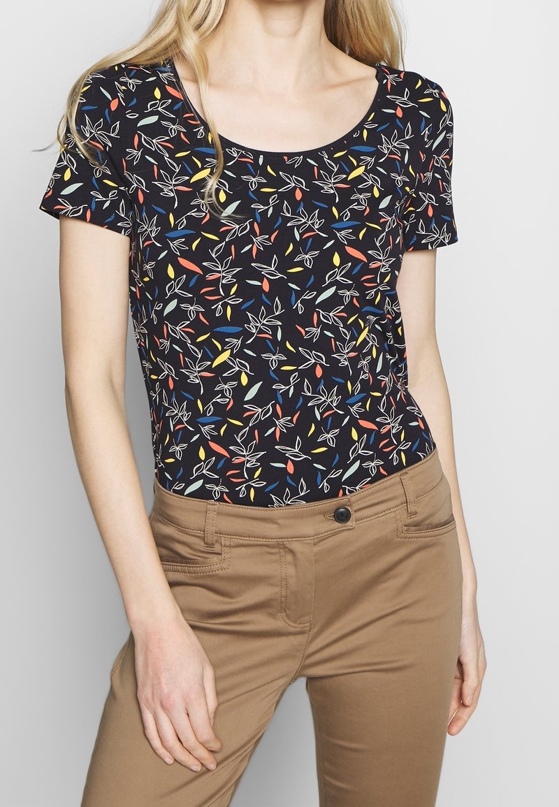 Black short-sleeve top with a colorful leaf pattern, featuring a scoop neckline. Paired with light brown fitted pants.