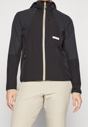 Giacca softshell nera con zip beige frontale completa e cappuccio, indossata con pantaloni beige chiaro, mani rilassate ai lati.