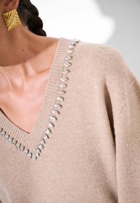Maglione beige a V con una texture aderente, decorato con accenti simili a gioielli trasparenti lungo il collo. Orecchini geometrici d'oro visibili.