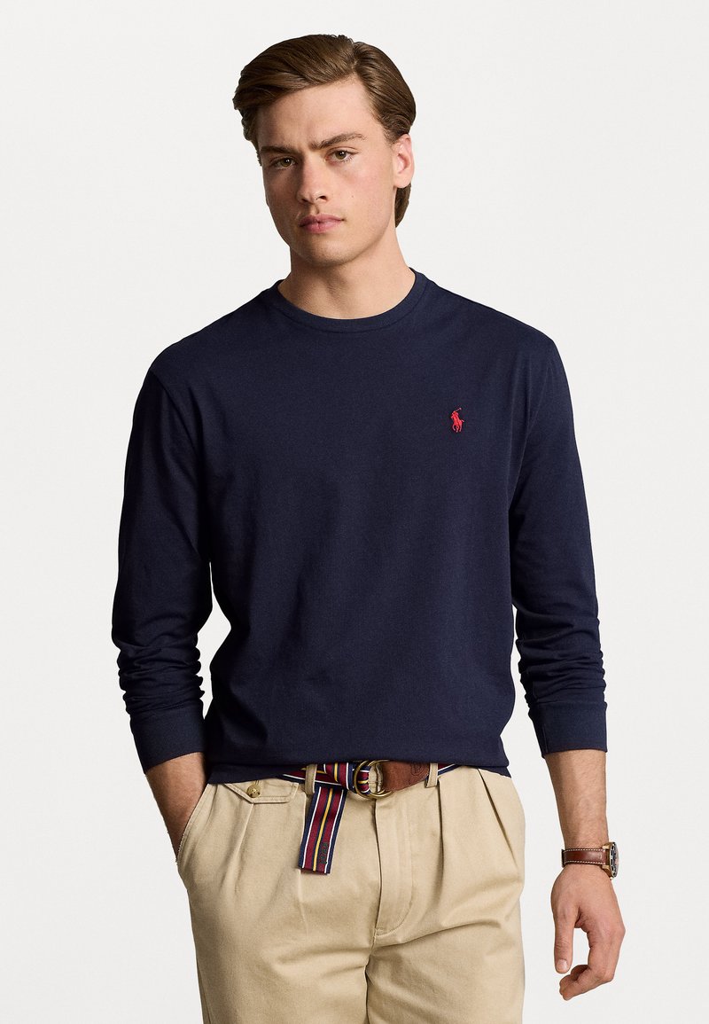 Marineblå langærmet t-shirt i bomuld, med et lille rødt logo på brystet. Båret med beige bukser og et flerfarvet stribet bælte.
