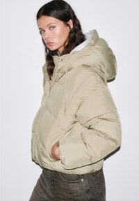 Giacca puffer khaki con cappuccio, caratterizzata da un design trapuntato, tasche frontali e polsini elasticizzati. Indossata sopra un indumento grigio.