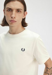 Camiseta de algodón crema, de mangas cortas, cuello redondo, con un logo de laurel bordado en azul marino en el pecho. Textura suave, ajuste casual.