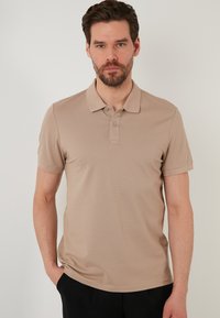 Bež polo majica s teksturiranim uzorkom, kratkim rukavima i ovratnikom na kojem se nalaze dva gumba, nosi se preko tamnih hlača.