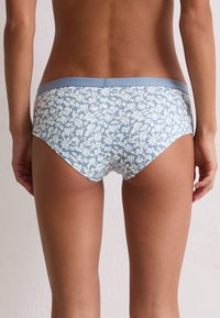 Vrouw in blauwe en witte bloemenmotief slips, gezien van achteren tegen een eenvoudige lichte muur als achtergrond.