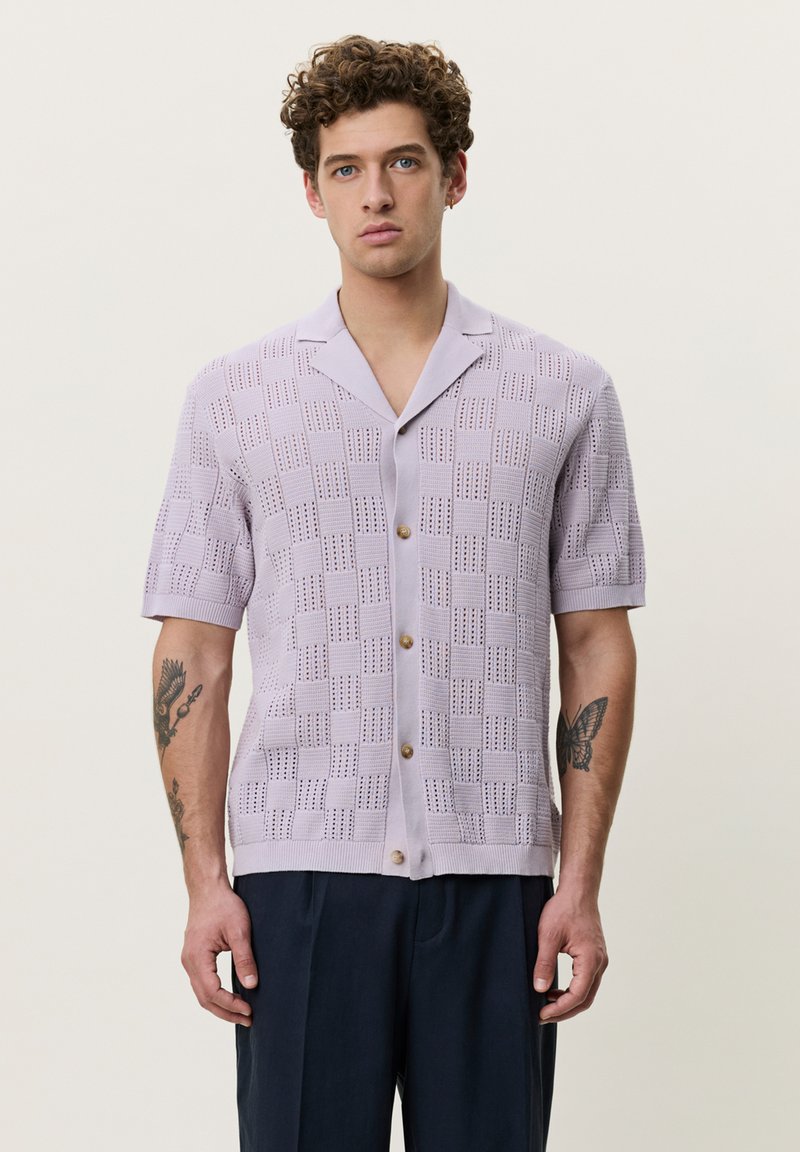 Jeune homme aux cheveux bouclés portant une chemise en maille à manches courtes violet clair avec un motif à carreaux et un pantalon foncé, debout devant un fond uni.