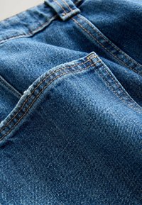 Denimstof in een middenblauwe kleur met een gestructureerd oppervlak. Kenmerkt zich door contrasterende oranje stiksels en rafelige randen bij de zoom.