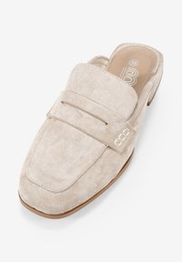 Zapato de ante beige sin cordones con punta redondeada, que presenta una correíta decorativa y un detalle de logotipo en relieve, complementado con un tacón bajo y sólido.