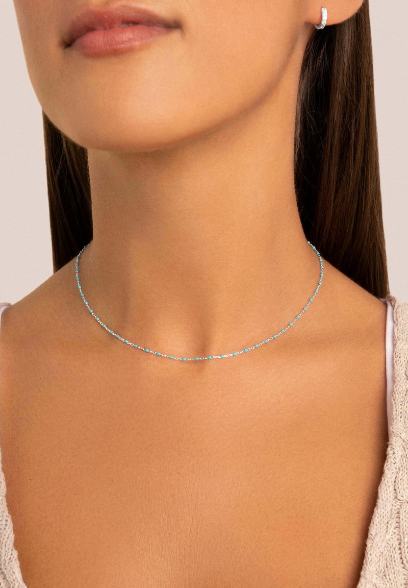 Collana blu sottile con piccoli anelli rotondi, poggiata sulla clavicola; abbinata a un orecchino a cerchio piccolo coordinato.