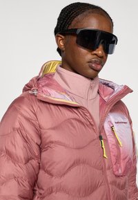 Ροζ φουσκωτό μπουφάν με υφή καπιτονέ, κίτρινη επένδυση και τσεπάκι με φερμουάρ. Φορεμένο πάνω από μπλούζα με ψηλό λαιμό; συνοδεύεται από μεγάλα μαύρα γυαλιά ηλίου.