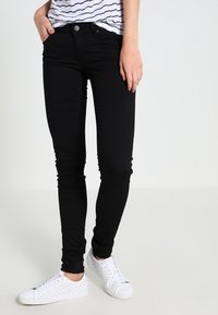 Vaqueros skinny negros de tejido elástico, con una cintura de altura media, bolsillos frontales y cierre con botón, combinados con zapatillas blancas.