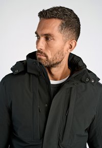 Svart, isolerad jacka med hög krage, frontdragkedja och två bröstfickor; har en slät, matt textur och minimal detaljer.