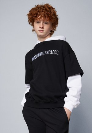 Sort sweatshirt med hvid tekst, lagd oven på en hvid langærmet skjorte. Har en afslappet pasform og ribbede manchetter, lavet af blødt materiale.