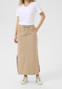 Jupe longue beige avec fentes latérales, taille élastique et cordon de serrage. Associée à un t-shirt blanc et des baskets de couleur claire.