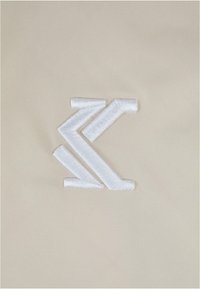 Beiger Stoff mit einem auffälligen weißen gestickten Logo, das aus kantigen Linien und Formen besteht, die symmetrisch angeordnet sind.