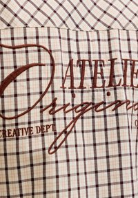 Καφέ κεντημένο κείμενο «Davelier Originals Creative Dept» σε φόντο υφάσματος με μπεζ και καφέ καρό σχέδιο.