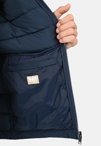 Intérieur d'une veste en duvet navy avec un rembourrage matelassé, fermeture éclair et une étiquette indiquant "T-KIT" sur un fond beige. Comprend une poche latérale.