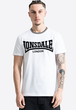 Mladý muž s vrkočmi nosí biele tričko Lonsdale London a ukazuje tetované pravé rameno s detailnými čiernymi dizajnmi z atramentu.