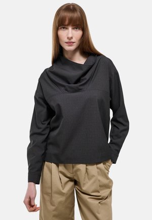 Femme portant un haut à manches longues gris foncé avec un col drapé et un pantalon beige plissé ample, debout devant un fond uni.