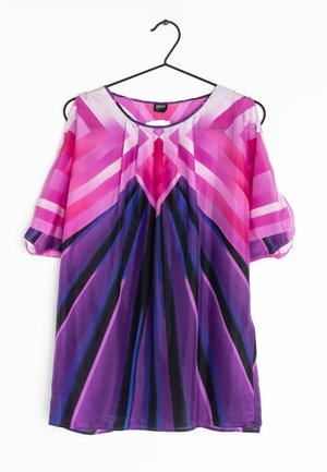Farverig, let bluse med lilla, lyserøde og sorte geometriske mønstre. Har brede ærmer og en rund halsudskæring. Hængende på en sort krog.
