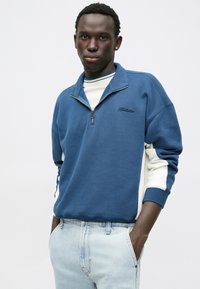 Sudadera azul con cremallera y paneles laterales blancos, con un cuello clásico y un logo de marca bordado en la parte frontal. Combinada con unos jeans de mezclilla claros.