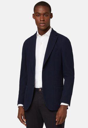 Man die een marineblauwe blazer draagt over een wit overhemd met knopen en een donkere broek, staand tegen een effen lichte achtergrond.