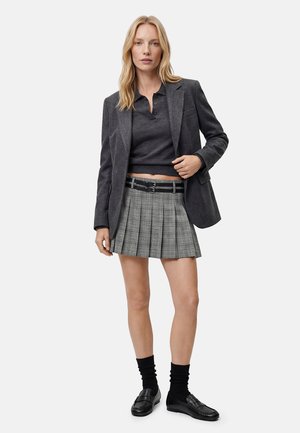 Kvinde stående iført en mørkegrå blazer, mørkegrå kort polo, grå ternet plisseret nederdel med bælte, sorte sokker og sorte loafers.