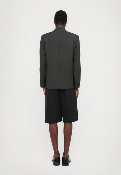 Γκρι κακοραμμένο σακάκι με λεπτομέρεια ραφής στο πίσω μέρος, συνδυασμένο με μαύρες βερμούδες, από την πίσω πλευρά. Μαύρα παπούτσια ολοκληρώνουν το σύνολο.