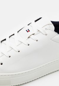 Sneaker in pelle bianca con lacci bianchi piatti, interno nero e un piccolo logo ricamato in rosso, bianco e blu sul lato.