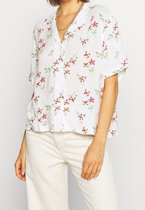 Vrouw draagt een wit kortärmlig overhemd met kleine roze bloemenprints en crèmegekleurde broek.