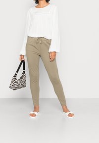 Camicia bianca leggera con dettagli plissettati, pantaloni aderenti verde oliva, sandali bianchi e borsa a righe bianca e nera.