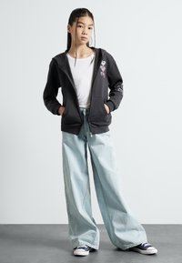 Zwarte zip-up hoodie met roze hart en tekstontwerp, gedragen over een wit t-shirt. Lichte blauwe wijde jeans en marineblauwe sneakers maken de outfit compleet.