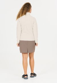 Kvinde med krøllet hår iført en lys beige langærmet top, brun nederdel og sorte sandaler, stående med ryggen mod på en hvid baggrund.