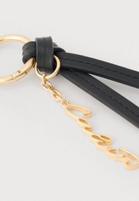 Porte-clés doré attaché à des lanières en cuir noir avec une chaîne dorée et un charm en or en forme de prénom en écriture cursive suspendu à l'anneau.